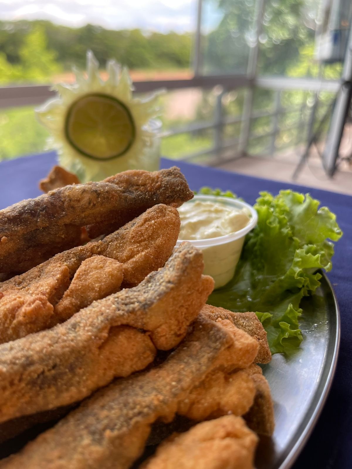 Peixe frito crocante servido com molho e folhas verdes, e uma caipirinha ao fundo